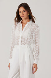 A woman is wearing a white sheer blouse with a floral pattern and a classic collar. The blouse is slightly transparent, revealing an underlying layer. She pairs it with high-waisted, wide-leg white trousers that have pleats at the front. She accessorizes with gold earrings and a ring on her right hand.